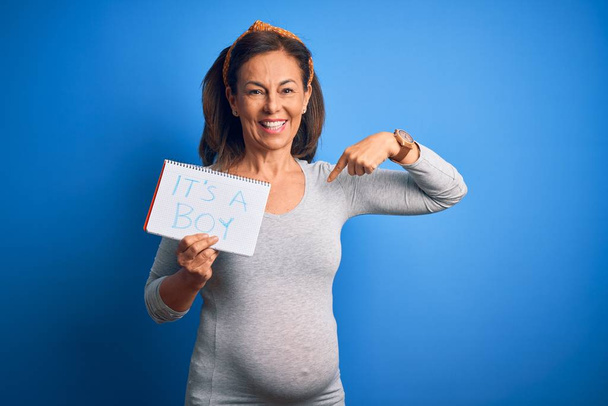 Orta yaşlı hamile bir kadın, mavi arka planda bir erkek bebek bekliyor. Suratı kendini işaret ediyor. - Fotoğraf, Görsel