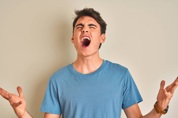 Tiener jongen draagt casual t-shirt staan over geïsoleerde achtergrond gek en gek schreeuwen en schreeuwen met agressieve uitdrukking en armen omhoog. Frustratieconcept. - Foto, afbeelding