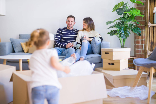 Güzel aile, anne-babalar kanepede kahve içme oturmuş karton kutular etrafında yeni evde oynayan çocuğuna bakıyor - Fotoğraf, Görsel