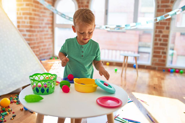 Anaokulunda bir sürü oyuncağın etrafında plastik yiyeceklerle oynayan sevimli sarışın çocuk. - Fotoğraf, Görsel