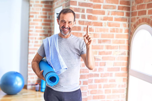 Middle age handsome sportman holding mat and towel standing before do exercise at gym surprised with an idea or question pointing finger with happy face, number one - Photo, image