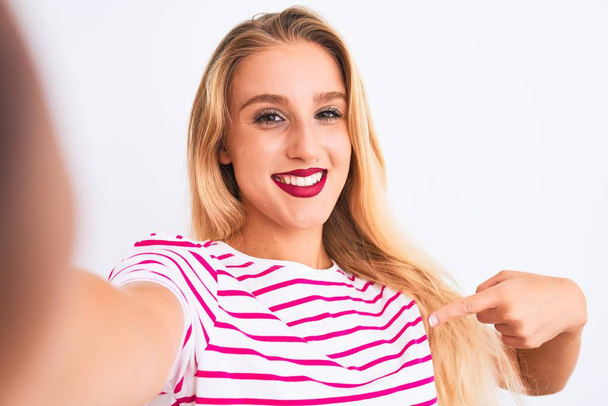 Beautiful woman wearing striped t-shirt make selfie by camera over isolated white background with surprise face pointing finger to himself - Photo, image