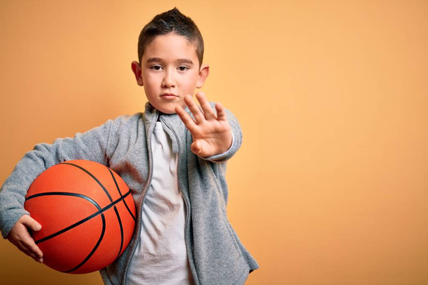 Nuori pieni poika poika leikkii koripallo peli pallo yli eristetty keltainen tausta avoimella kädellä tekee stop merkki vakava ja luottavainen ilme, puolustus ele
 - Valokuva, kuva