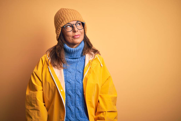 Mujer de mediana edad vistiendo impermeable amarillo y sombrero de invierno sobre fondo aislado sonriendo mirando hacia un lado y mirando hacia otro pensando
. - Foto, Imagen