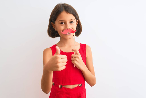 Beautiful child girl holding fanny party mustache standing over isolated white background happy with big smile doing ok sign, thumb up with fingers, excellent sign - Photo, Image