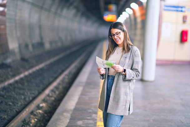 Молодая красивая женщина улыбается счастливой и уверенной. Стоя с улыбкой на лице, держа билеты на платформе железнодорожного вокзала
 - Фото, изображение