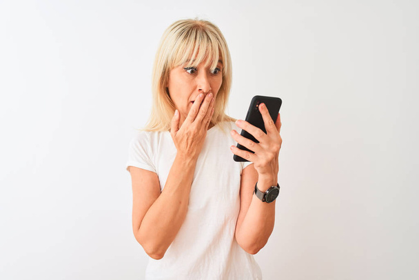 Middle age woman using smartphone standing over isolated white background cover mouth with hand shocked with shame for mistake, expression of fear, scared in silence, secret concept - Foto, Imagem
