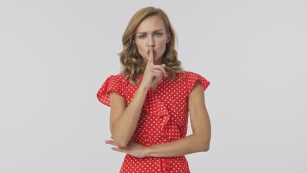 Serious pretty blonde woman in dress showing silence gesture and looking at the camera while standing with crossed arms over grey background - Felvétel, videó