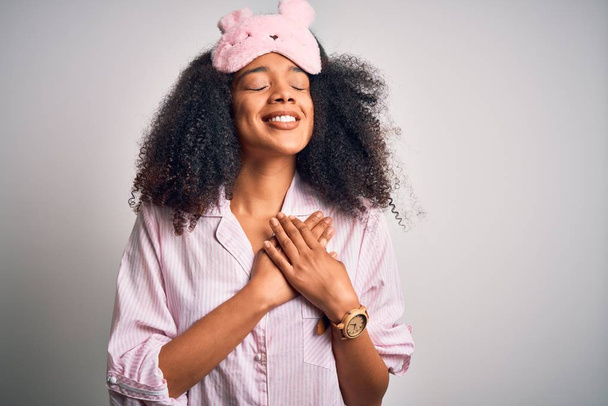 Mujer afroamericana joven con cabello afro con máscara para dormir y pijama rosa sonriendo con las manos en el pecho con los ojos cerrados y gesto agradecido en la cara. Concepto de salud
. - Foto, Imagen