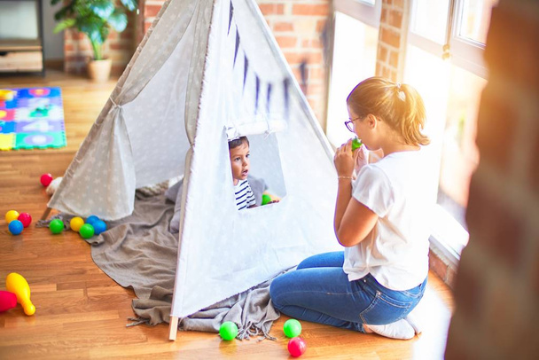 Beautiful teacher and toddler boy sitting on the floor playing inside  tipi at kindergarten - Photo, Image