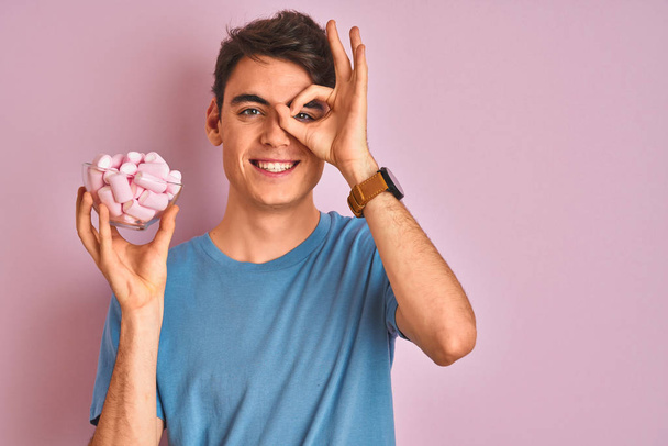 Teenager boy holding a bunch of delicious fluffy marshmallow over isolated pink background with happy face smiling doing ok sign with hand on eye looking through fingers - Photo, Image