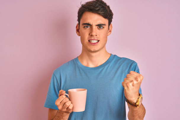 Teenager Junge, der eine Tasse Kaffee vor isoliertem rosa Hintergrund trinkt, genervt und frustriert schreit vor Wut, verrückt und schreit mit erhobener Hand, Wut-Konzept - Foto, Bild