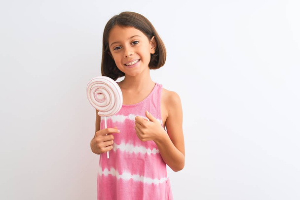 Beautiful child girl eating sweet lollipop standing over isolated white background happy with big smile doing ok sign, thumb up with fingers, excellent sign - Foto, immagini