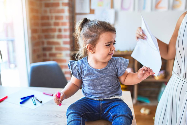 Hermoso niño sentado en el escritorio llorando y joven profesor mostrando dibujo en el jardín de infantes
 - Foto, Imagen