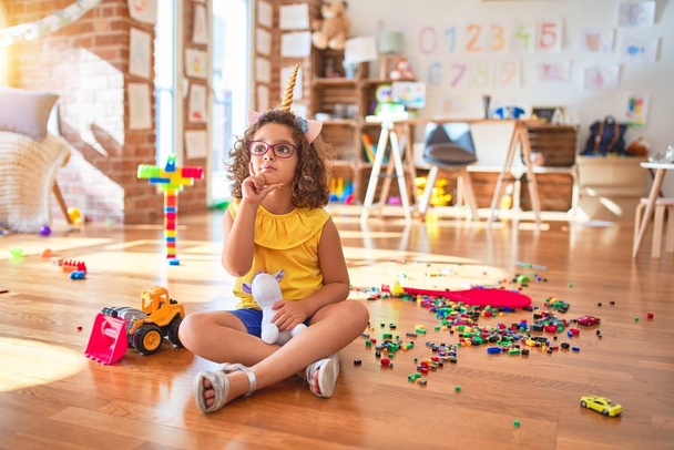 Beautiful toddler wearing glasses and unicorn diadem sitting on the floor at kindergarten with hand on chin thinking about question, pensive expression. Smiling with thoughtful face. Doubt concept. - Photo, image