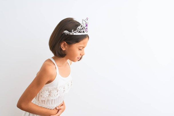 Gyönyörű gyermek lány visel hercegnő koronát áll felett elszigetelt fehér háttér kézzel a gyomorban, mert emésztési zavar, fájdalmas betegség rossz közérzet. Ache-koncepció. - Fotó, kép