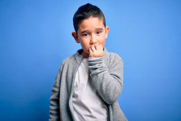 Młody chłopiec noszący sportową bluzę na niebieskim, odizolowanym tle wyglądający na zestresowanego i nerwowego z rękami obgryzającymi paznokcie. Problem lękowy. - Zdjęcie, obraz