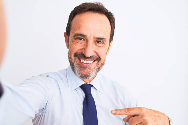 Hombre de negocios de mediana edad con corbata hacer selfie de pie sobre fondo blanco aislado muy feliz señalando con la mano y el dedo
 - Foto, imagen