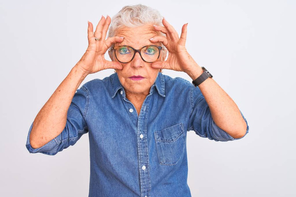 Femme âgée aux cheveux gris portant une chemise en denim et des lunettes sur fond blanc isolé Essayant d'ouvrir les yeux avec les doigts, somnolente et fatiguée pour la fatigue matinale
 - Photo, image