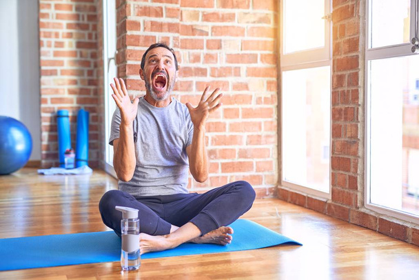 gutaussehender Sportler mittleren Alters, der mit erhobenen Armen und offenen Augen aufgeregt auf einer Matte sitzt und Stretching-Yoga-Übungen im Fitnessstudio macht, feiert verrückt und erstaunt über den Erfolg. Siegerkonzept - Foto, Bild