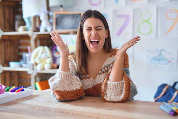 Young beautiful teacher woman wearing sweater and glasses sitting on desk at kindergarten celebrating mad and crazy for success with arms raised and closed eyes screaming excited. Winner concept - Valokuva, kuva