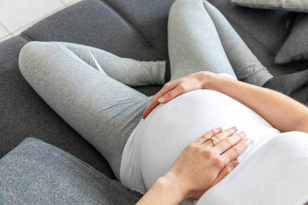 Fiatal terhes nő tartja a hasát ül egy kanapén - Fotó, kép