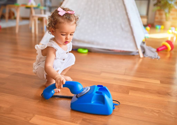 Beautiful caucasian infant playing with toys at colorful playroom. Happy and playful calling with vitange telephone at kindergarten. - 写真・画像