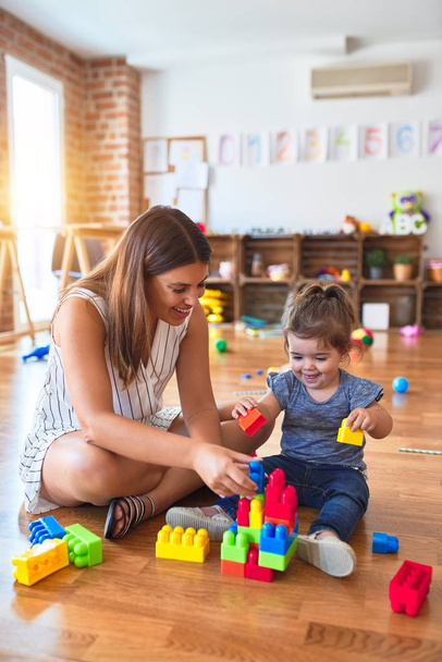 Young beautiful teacher and toddler playing with building blocks toy at kindergarten - 写真・画像