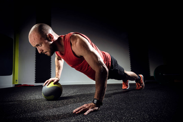 Muscular young man doing push ups with medicine ball on dark studio. - Фото, изображение