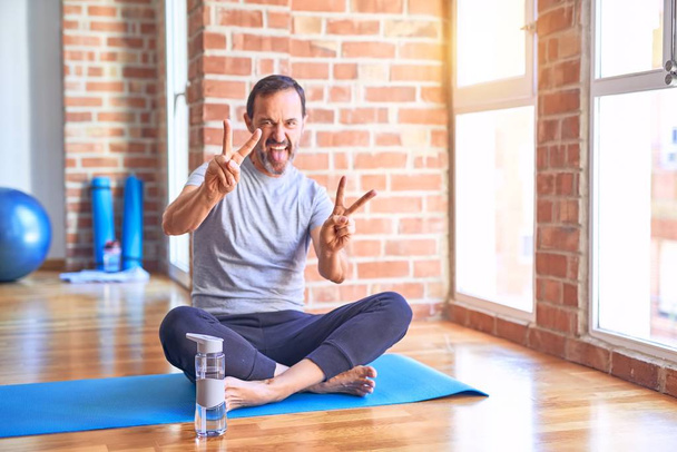 Średniego wieku przystojny sportowiec siedzi na macie robiąc rozciągające ćwiczenia jogi na siłowni uśmiechając się z językiem na zewnątrz pokazując palce obu rąk robi znak zwycięstwa. Numer dwa.. - Zdjęcie, obraz