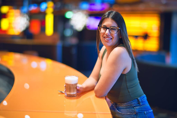 Молодая красивая женщина улыбается счастливой и уверенной. Опираясь на стойку бара держа стакан кофе в ресторане
 - Фото, изображение