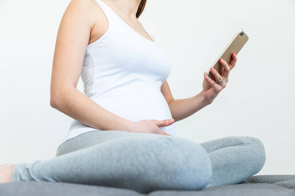 Young pregnant woman holds her belly and looks at her phone whil - 写真・画像