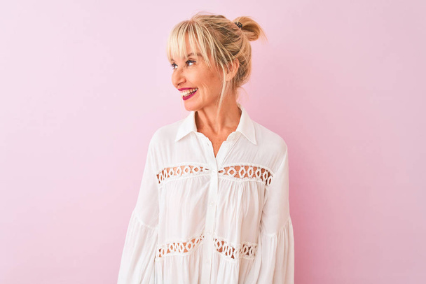 Middle age woman wearing white casual shirt standing over isolated pink background looking away to side with smile on face, natural expression. Laughing confident. - Photo, Image