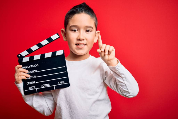 Маленький мальчик, снимающий видео, держа режиссера кинотеатра на изолированном красном фоне, удивленный идеей или вопросом, указывающим пальцем со счастливым лицом, номер один
 - Фото, изображение