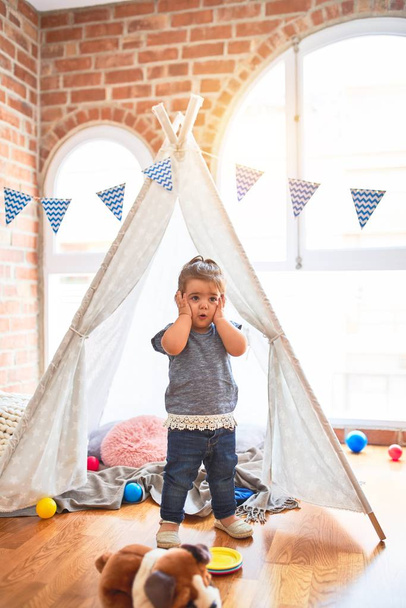 Beautiful toddler standing inside tipi with hands on face surprised and worried at kindergarten - Photo, image