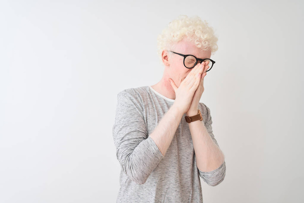 junger Albino-blonder Mann mit gestreiftem T-Shirt und Brille über isoliertem weißem Hintergrund, der etwas Stinkendes und ekelhaften, unerträglichen Geruch riecht und mit den Fingern auf der Nase den Atem anhält. Konzept für schlechte Gerüche. - Foto, Bild