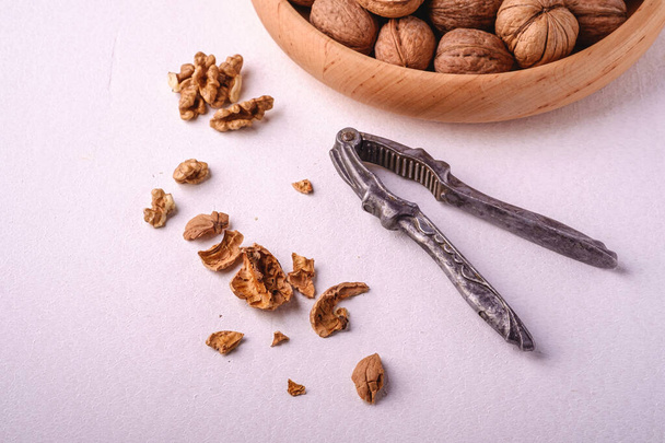Walnuts heap food in wooden bowl with half peeled nut, cracked nutshell, near to vintage nutcracker on white background, angle view, healthy food concept - Photo, image