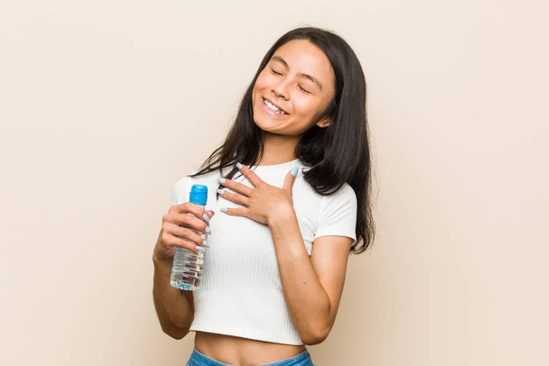 młoda Azjatka trzyma butelkę wody śmieje się głośno trzymając rękę na klatce piersiowej. - Zdjęcie, obraz