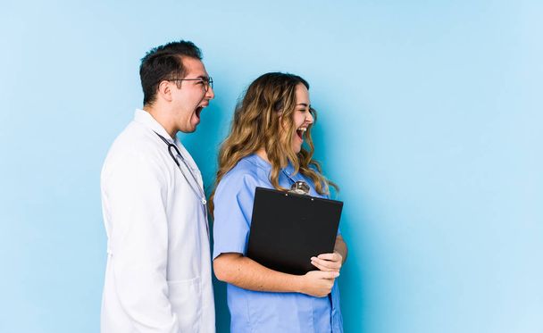 Joven pareja de médicos posando en un fondo azul aislado gritando hacia un espacio de copia
 - Foto, Imagen