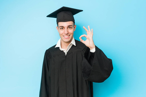 Молодой кавказский выпускник мужчина веселый и уверенный в себе показывая ОК жест
. - Фото, изображение