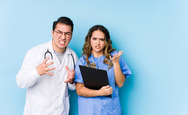 joven doctor pareja posando en un azul fondo aislado molesto gritando con tensas manos
. - Foto, Imagen