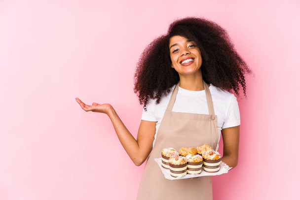 Jonge afro gebak maker vrouw met een cupcakes geïsoleerde jonge afro bakker vrouw tonen een kopie ruimte op een palm en houden een andere hand op taille. - Foto, afbeelding
