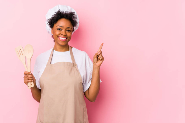 Mladá africká americká kuchařka se usmívá a ukazuje stranou, ukazuje něco na prázdném místě. - Fotografie, Obrázek