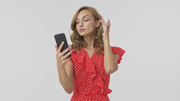 Calm pretty blonde woman in dress using smartphone like a mirror and smartening over grey background - Imágenes, Vídeo