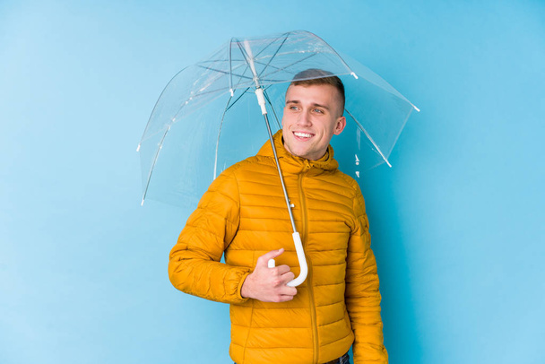 Joven hombre caucásico sosteniendo un paraguas mira a un lado sonriente, alegre y agradable
. - Foto, imagen