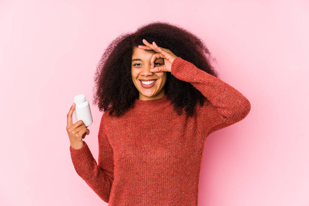 Νεαρή Αφρο γυναίκα που κρατάει τις βιταμίνες απομονωμένες Νεαρή Αφρο γυναίκα που κρατάει τις βιταμίνες της ενθουσιασμένοι κρατώντας την οκέι χειρονομία στο μάτι. - Φωτογραφία, εικόνα