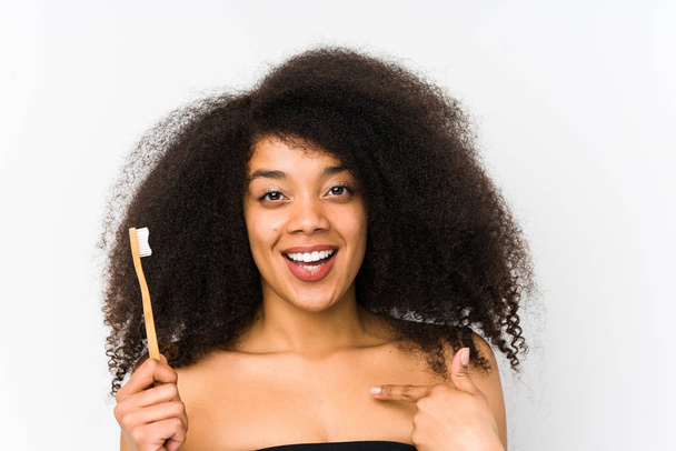 Νεαρή Αφρο γυναίκα κρατώντας μια οδοντόβουρτσα απομονωμένη έκπληκτη δείχνοντας τον εαυτό του, χαμογελώντας ευρέως. - Φωτογραφία, εικόνα