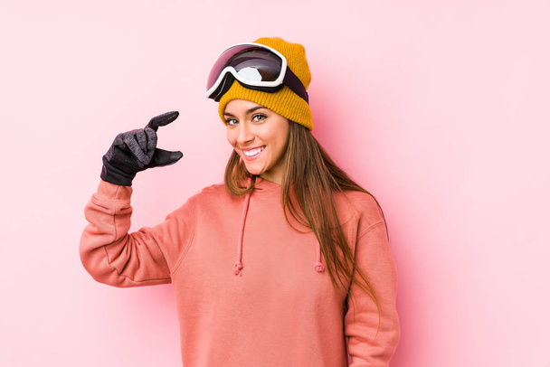 Νεαρή λευκή γυναίκα που φοράει ρούχα του σκι απομονωμένη κρατώντας κάτι μικρό με τα μπροστινά δάχτυλα, χαμογελαστή και σίγουρη. - Φωτογραφία, εικόνα
