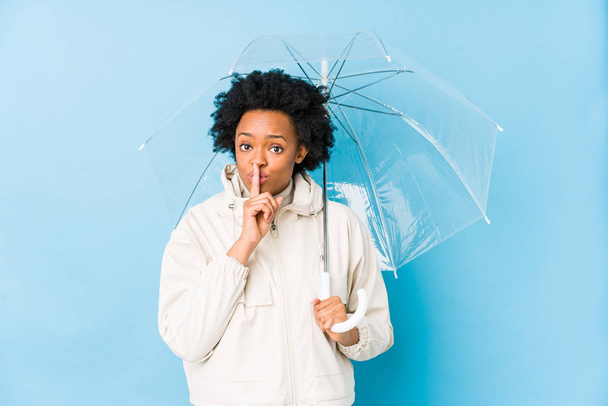 Joven mujer afroamericana sosteniendo un paraguas aislado guardando un secreto o pidiendo silencio
. - Foto, Imagen
