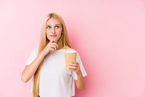 junges blondes Mädchen mit einem Kaffee zum Mitnehmen, das mit zweifelndem und skeptischem Gesichtsausdruck zur Seite schaut. - Foto, Bild